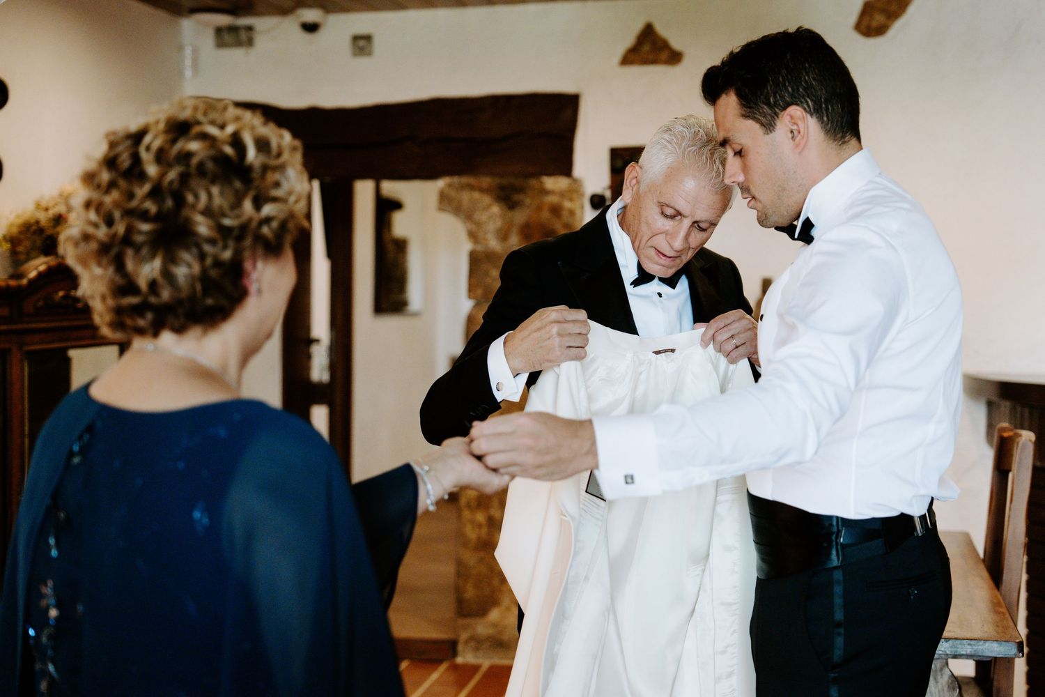 Groom being helped into white tuxedo jacket by two family members before wedding ceremony indoors.