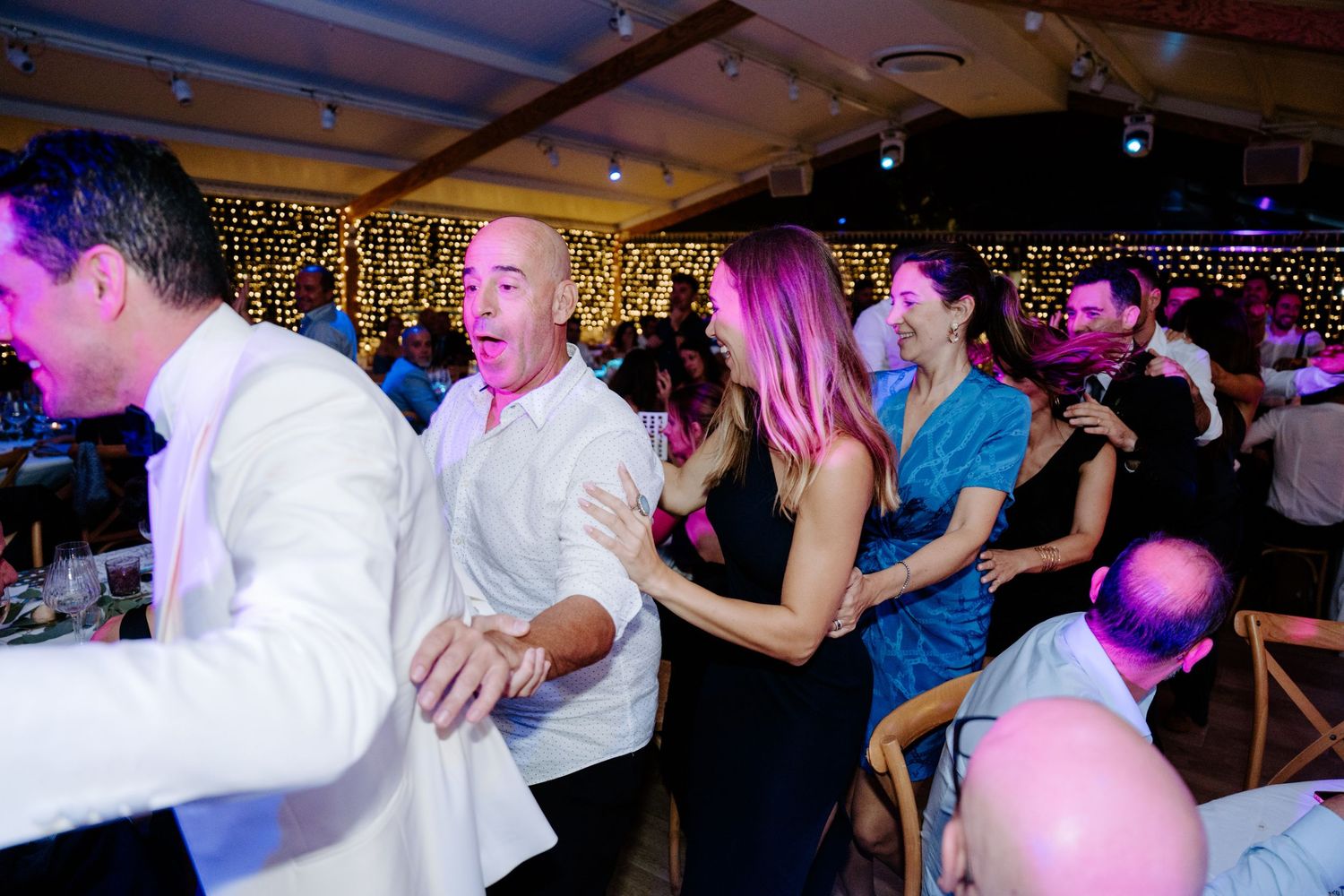 Guests dancing and celebrating at a lively wedding reception under warm fairy lights.
