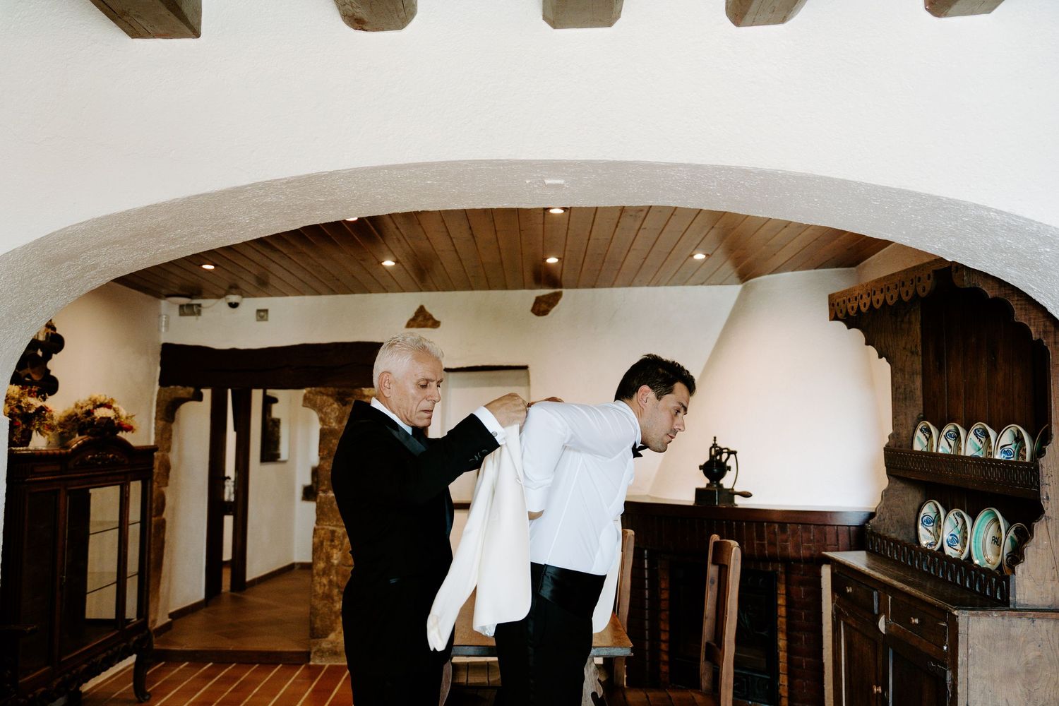 Older man helping groom put on white wedding jacket in rustic interior.
