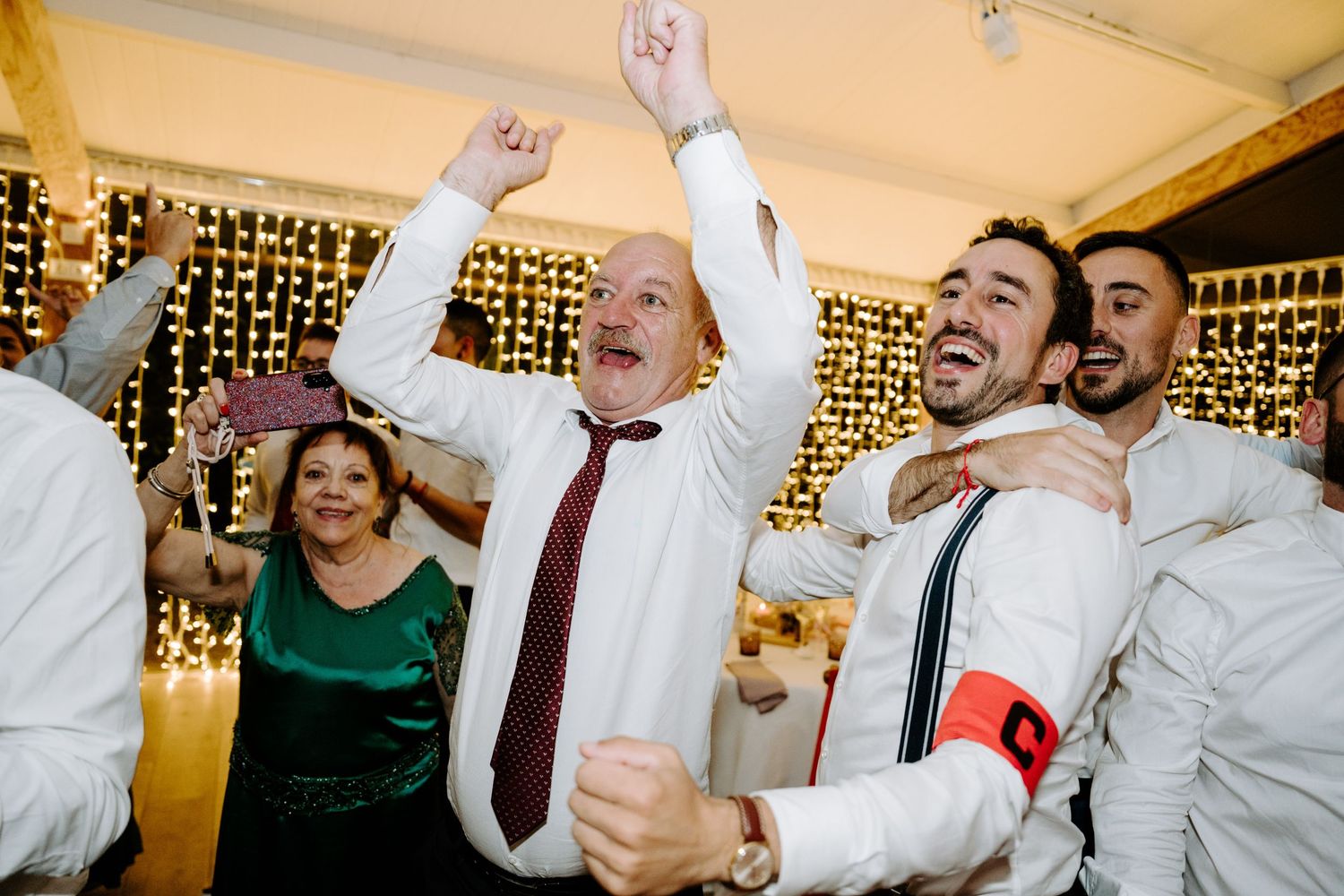 Joyful guests celebrating and dancing at a wedding reception with fairy lights in the background.