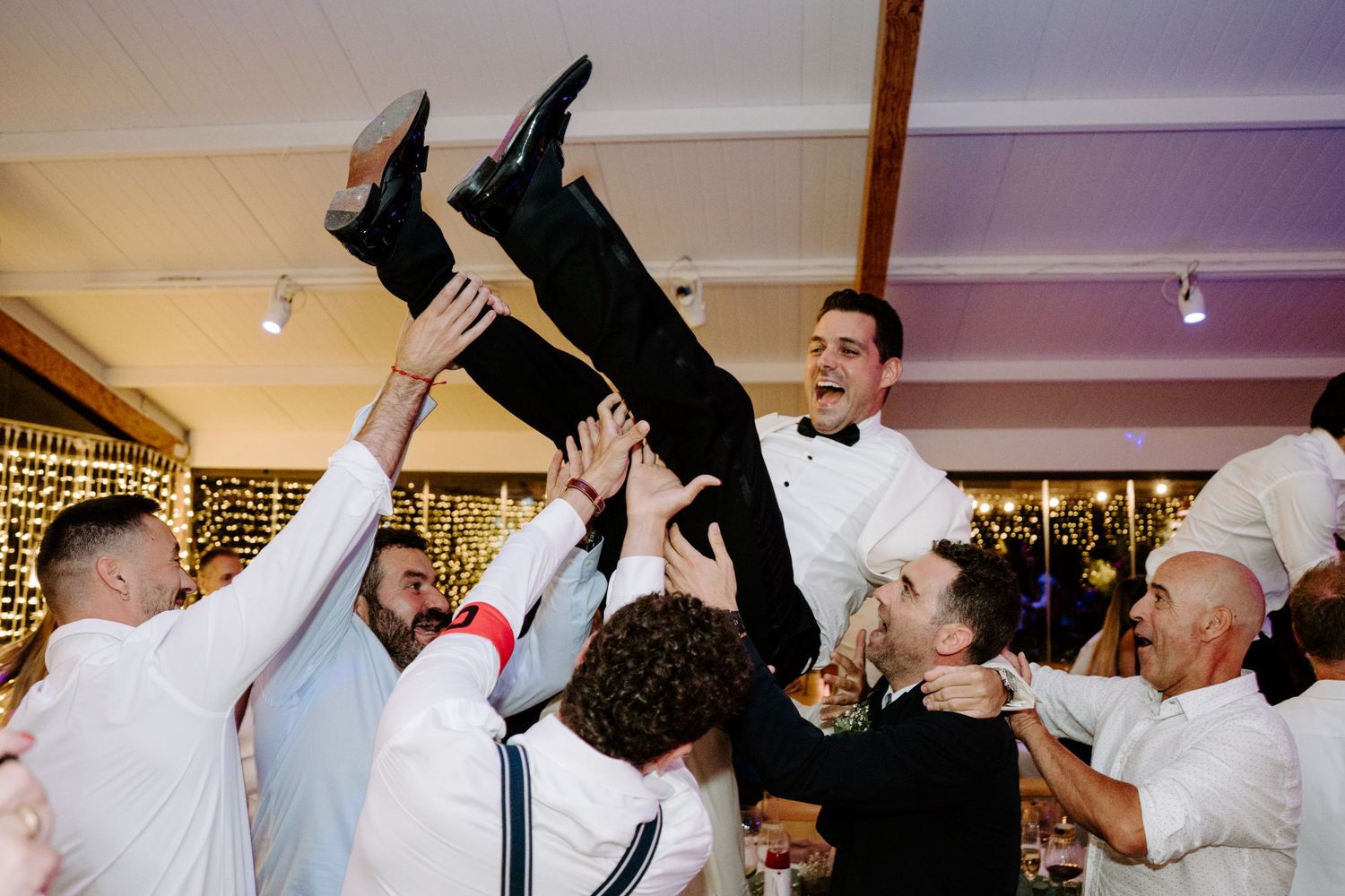 Groomsmen lifting groom in the air during wedding reception celebration.
