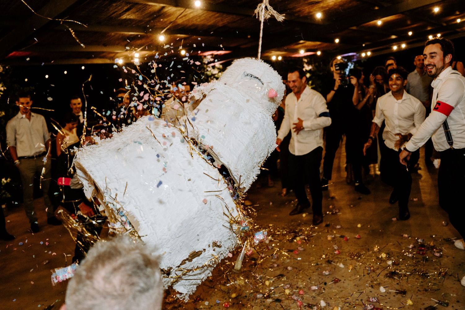 Guests smashing a white piñata with confetti exploding at a lively wedding reception party.