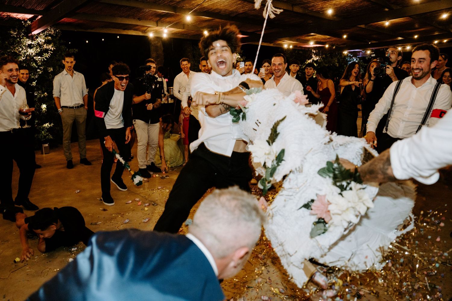 Groom smashing wedding cake surrounded by cheering guests at reception.