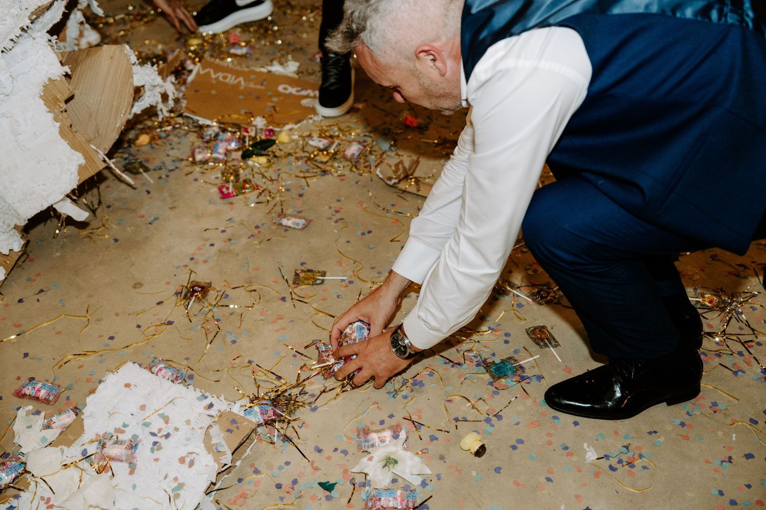 Man in white shirt and blue vest crouching to pick up colorful confetti from a debris-covered floor at a wedding reception.