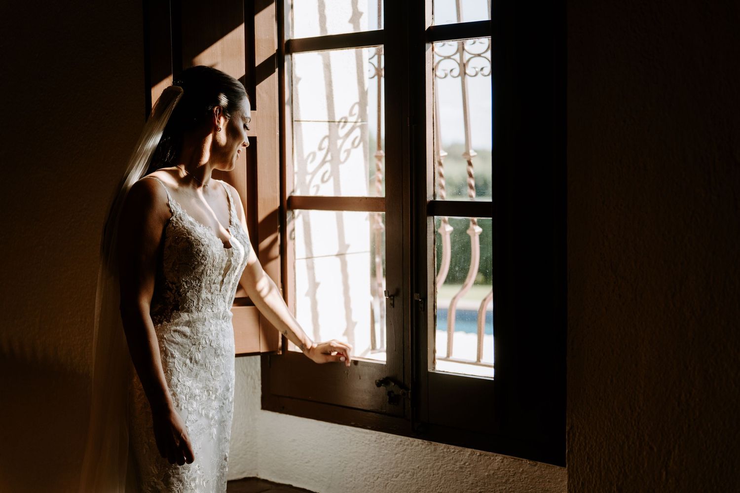 Bride in lace wedding gown stands by ornate wooden window in soft natural light.