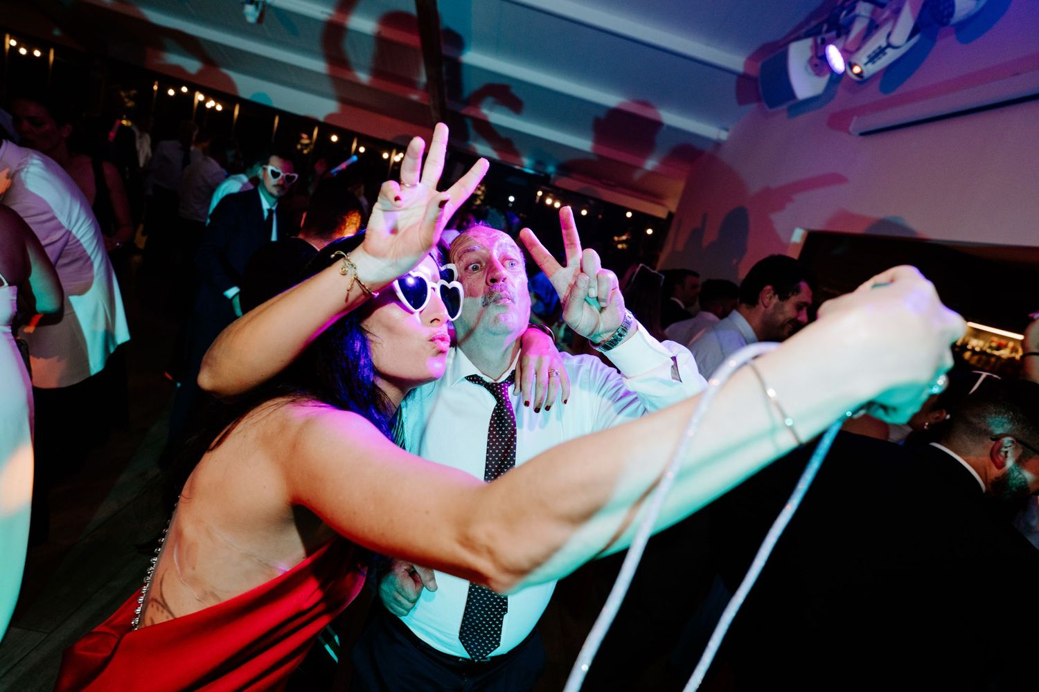 Guests dancing at a lively wedding reception party wearing fun white sunglasses under colorful lights.