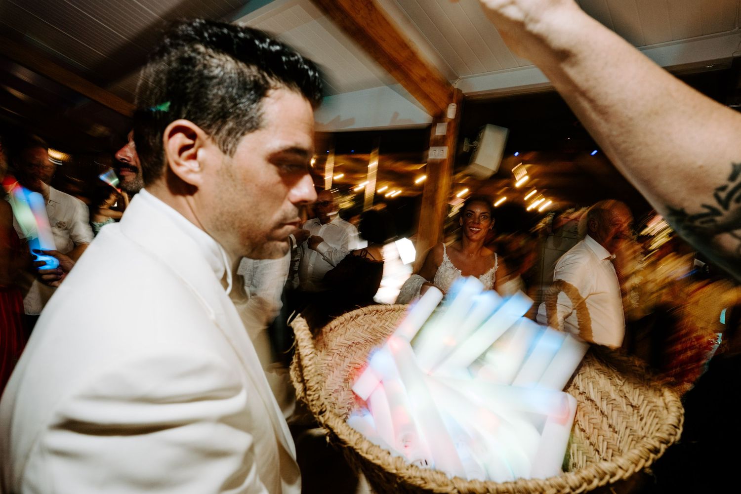 Groom in white jacket selecting a glowing light stick from a wicker basket at a lively wedding reception party.
