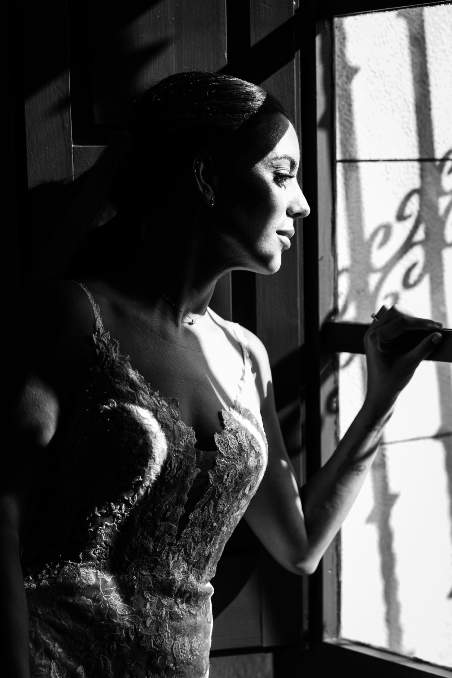 Elegant bride in lace gown gazing out window in black and white portrait.