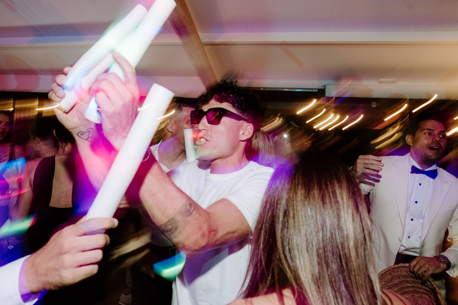 Man in white shirt and sunglasses dancing with glow sticks at a lively wedding party.