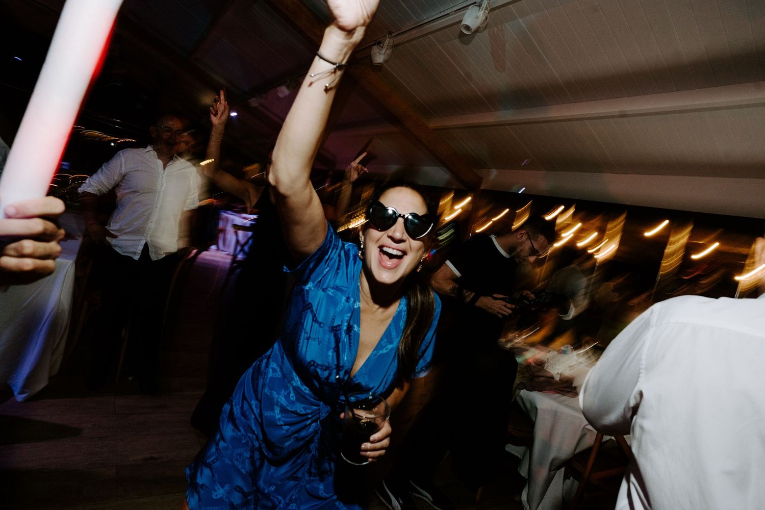 Excited wedding guest in blue dress and sunglasses dancing with a glow stick at reception.