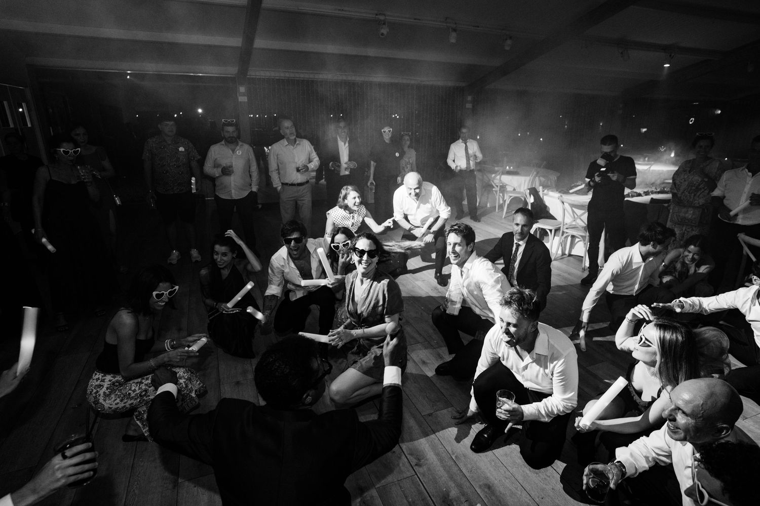 Black and white photo of wedding guests sitting and dancing on the floor at a lively indoor wedding reception.