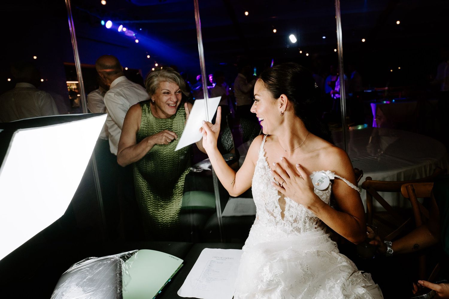 Bride in white lace gown reacting with joy while opening a gift at her wedding reception with a smiling guest nearby.