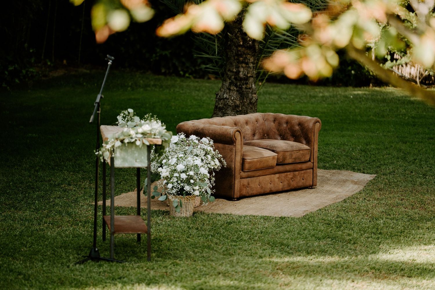 Rustic leather Chesterfield sofa on a jute rug surrounded by white floral arrangements in an outdoor garden wedding ceremony.
