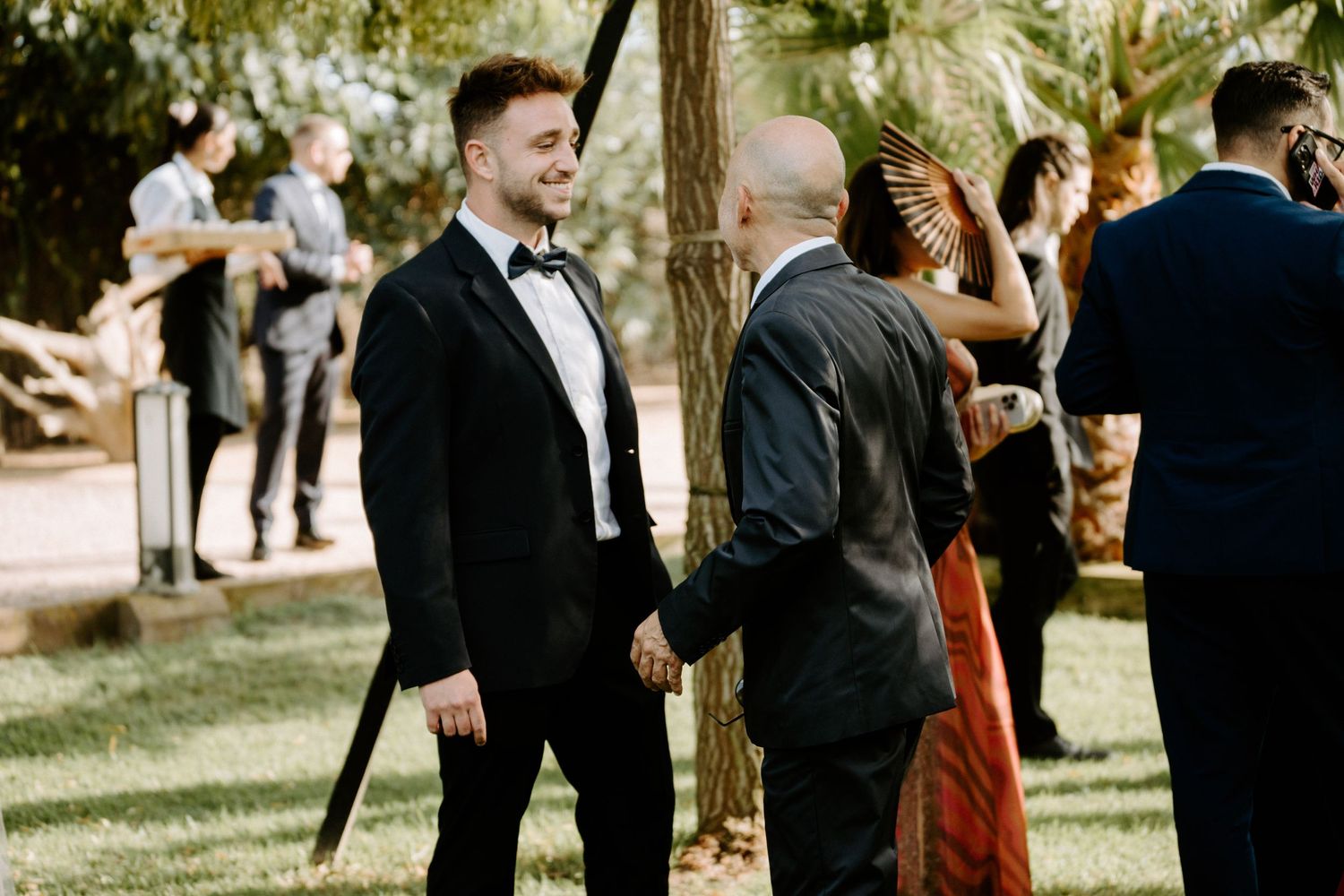 Smiling groom in black tuxedo greeting bald guest outdoors at wedding cocktail hour.