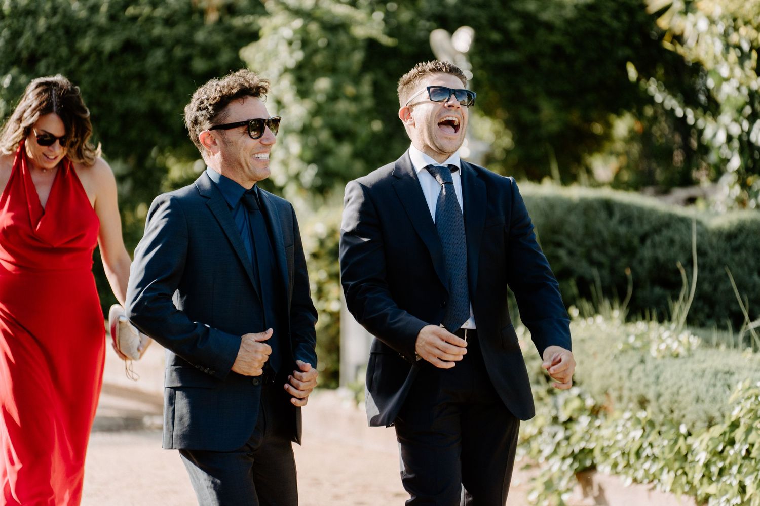 Two men in navy suits and sunglasses laughing while walking through a garden wedding venue.
