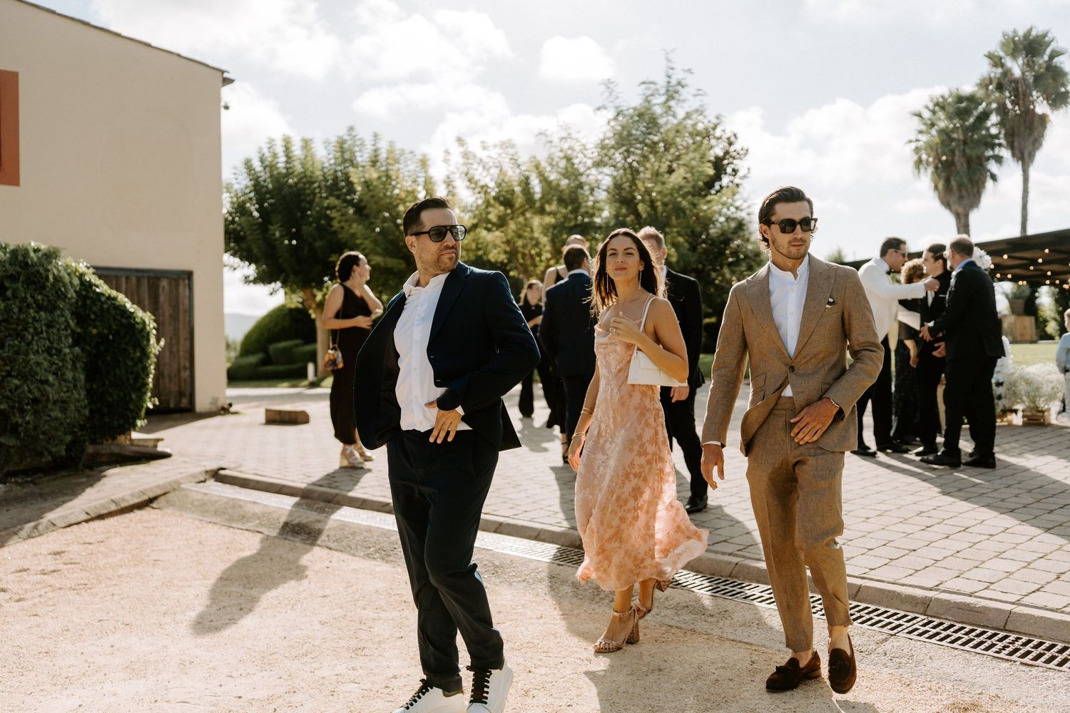 Three elegantly dressed wedding guests walking together at sunny outdoor venue.