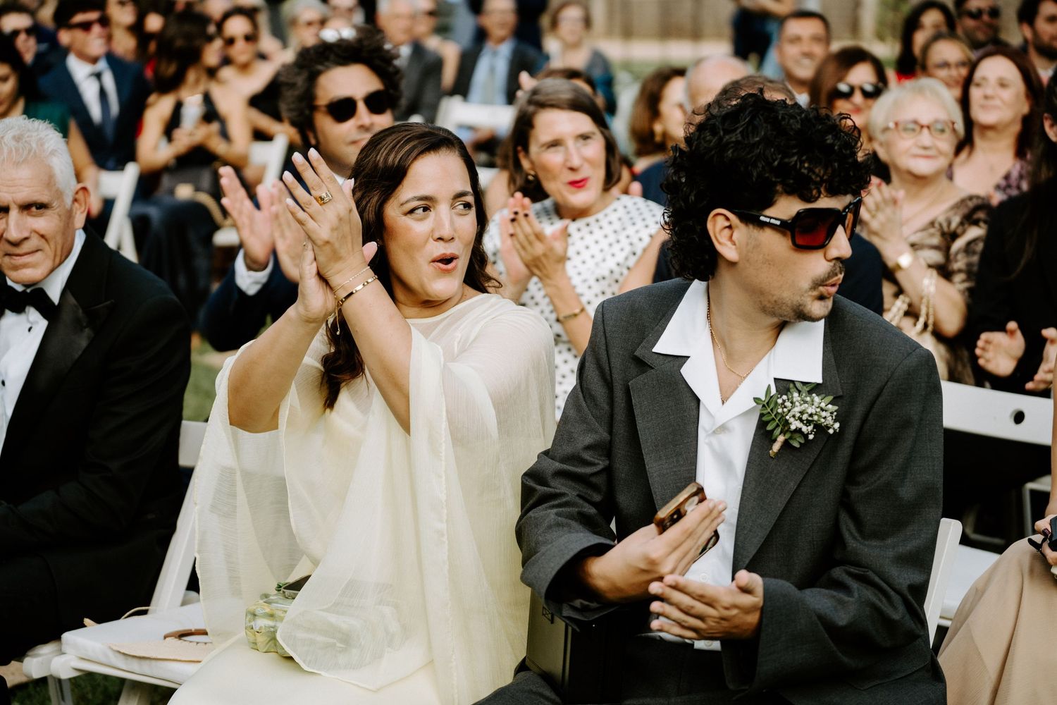 Wedding guests clap and react with excitement during outdoor daytime wedding ceremony seating.