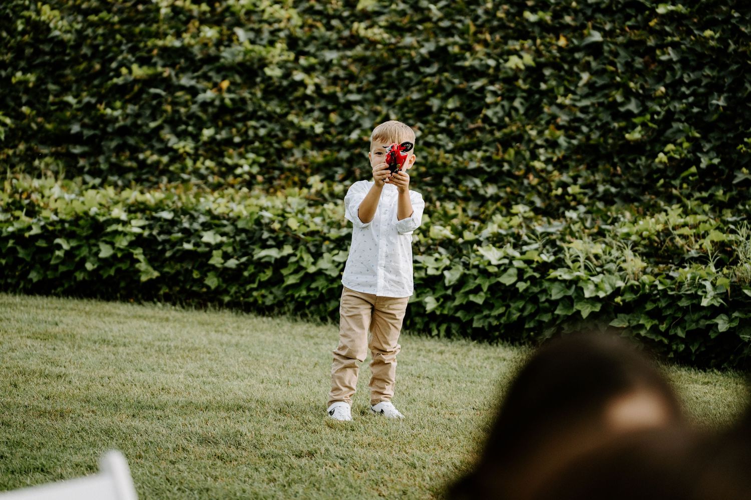 Young child in casual clothes playing with a red toy on a lush green lawn at a wedding.