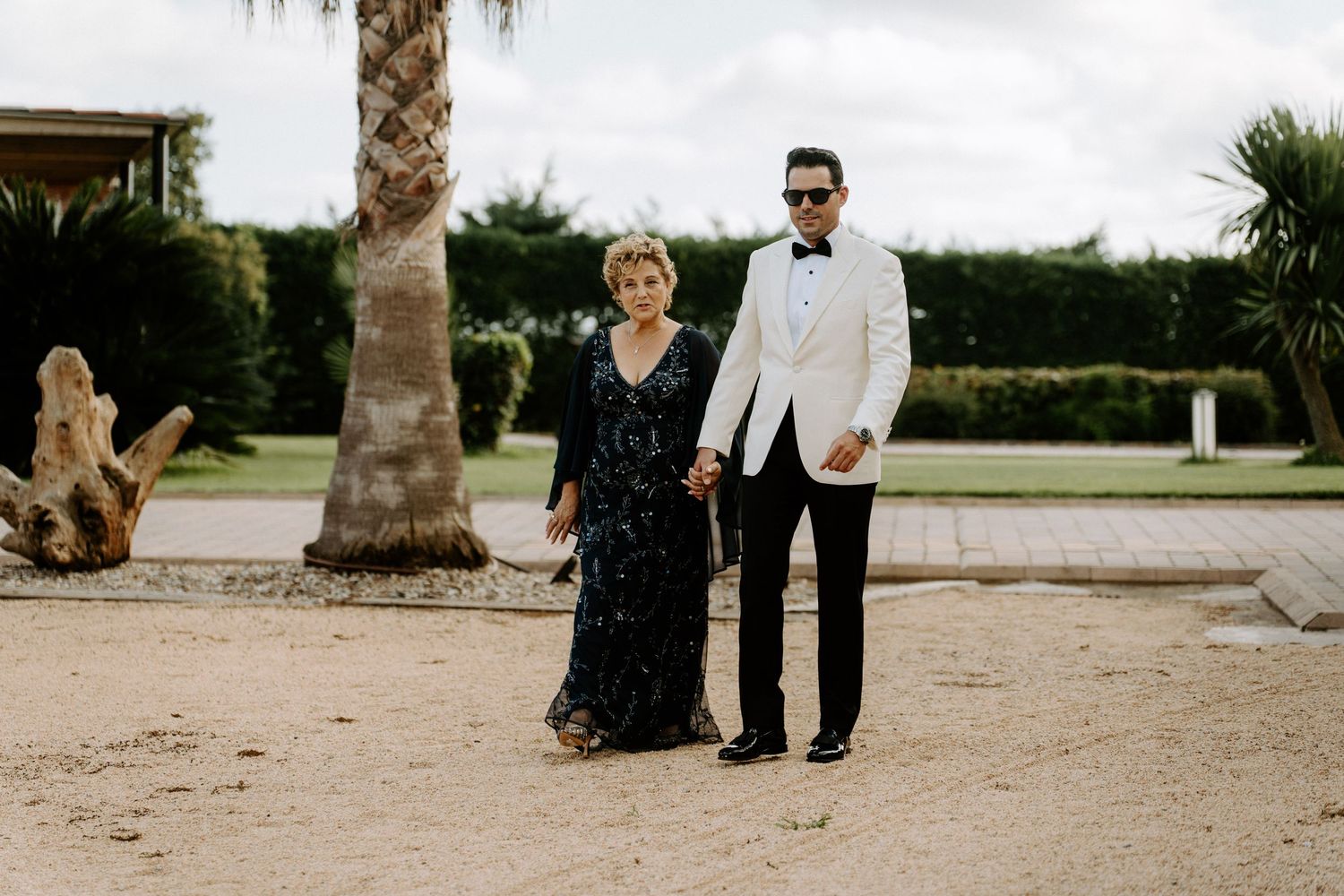 Groom in white tuxedo holds hands with elegantly dressed woman outdoors at wedding venue.