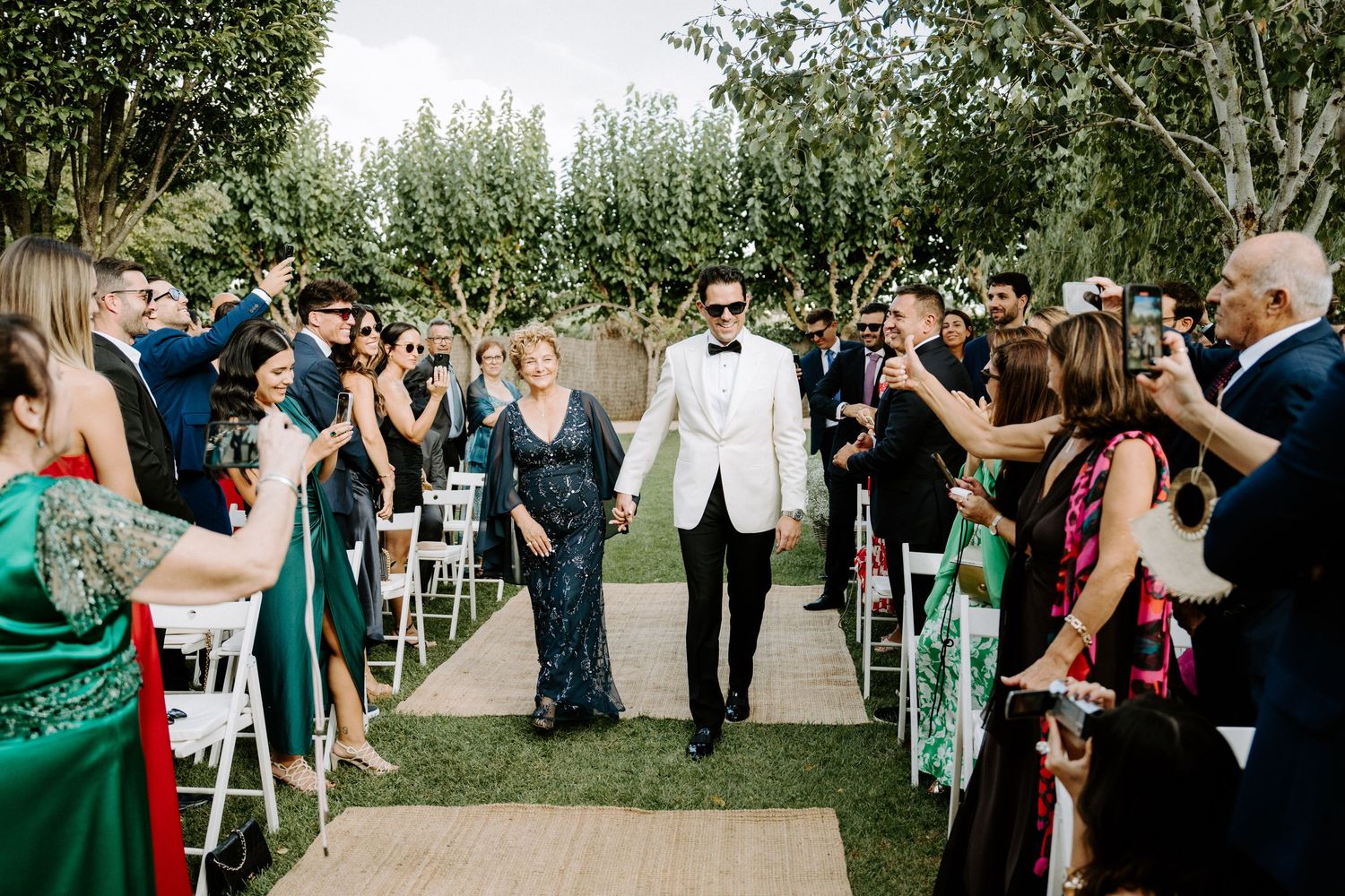 Groom walking down outdoor aisle with mother during sunny garden wedding ceremony.