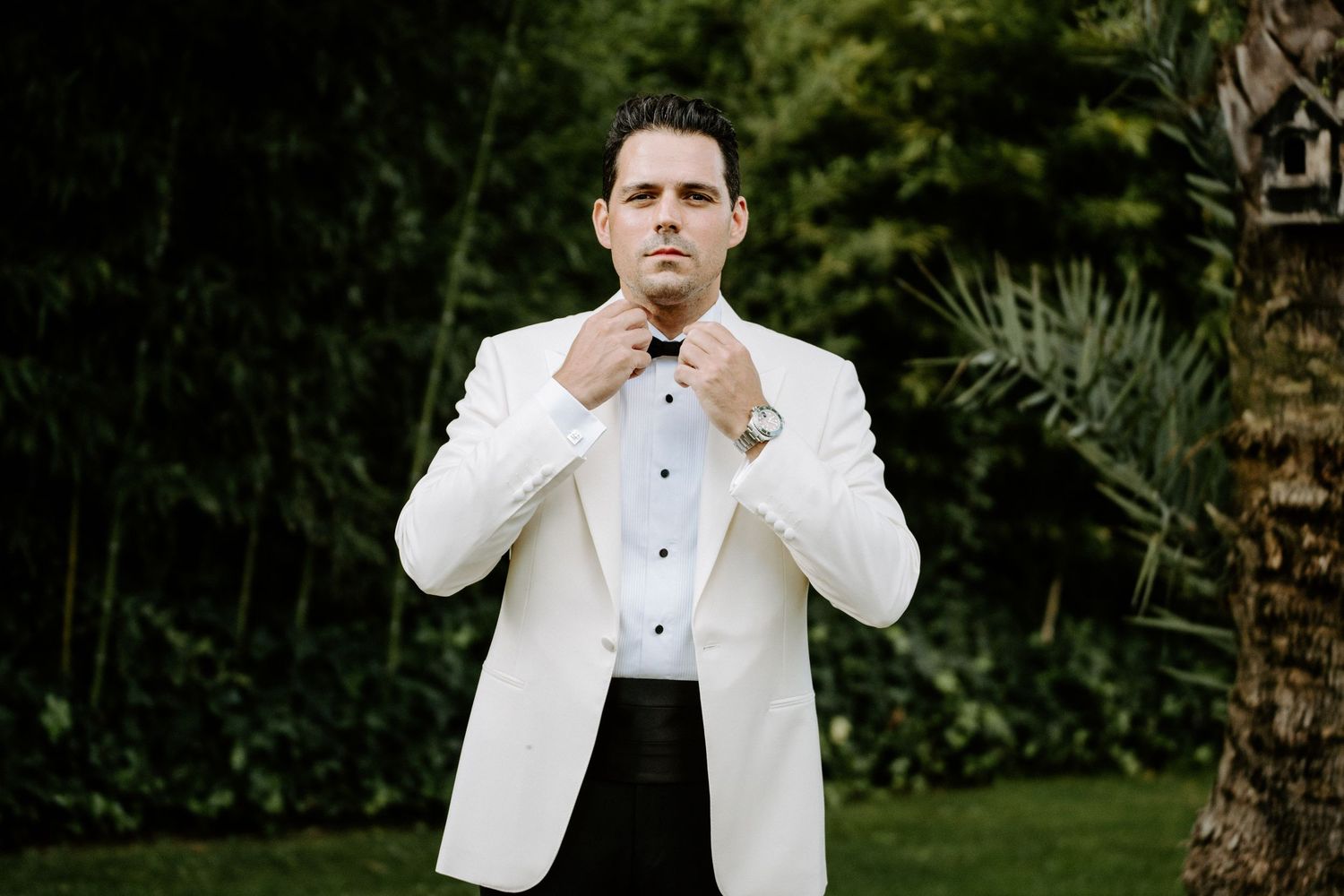 Groom in white tuxedo jacket adjusting bow tie outdoors in lush garden setting.