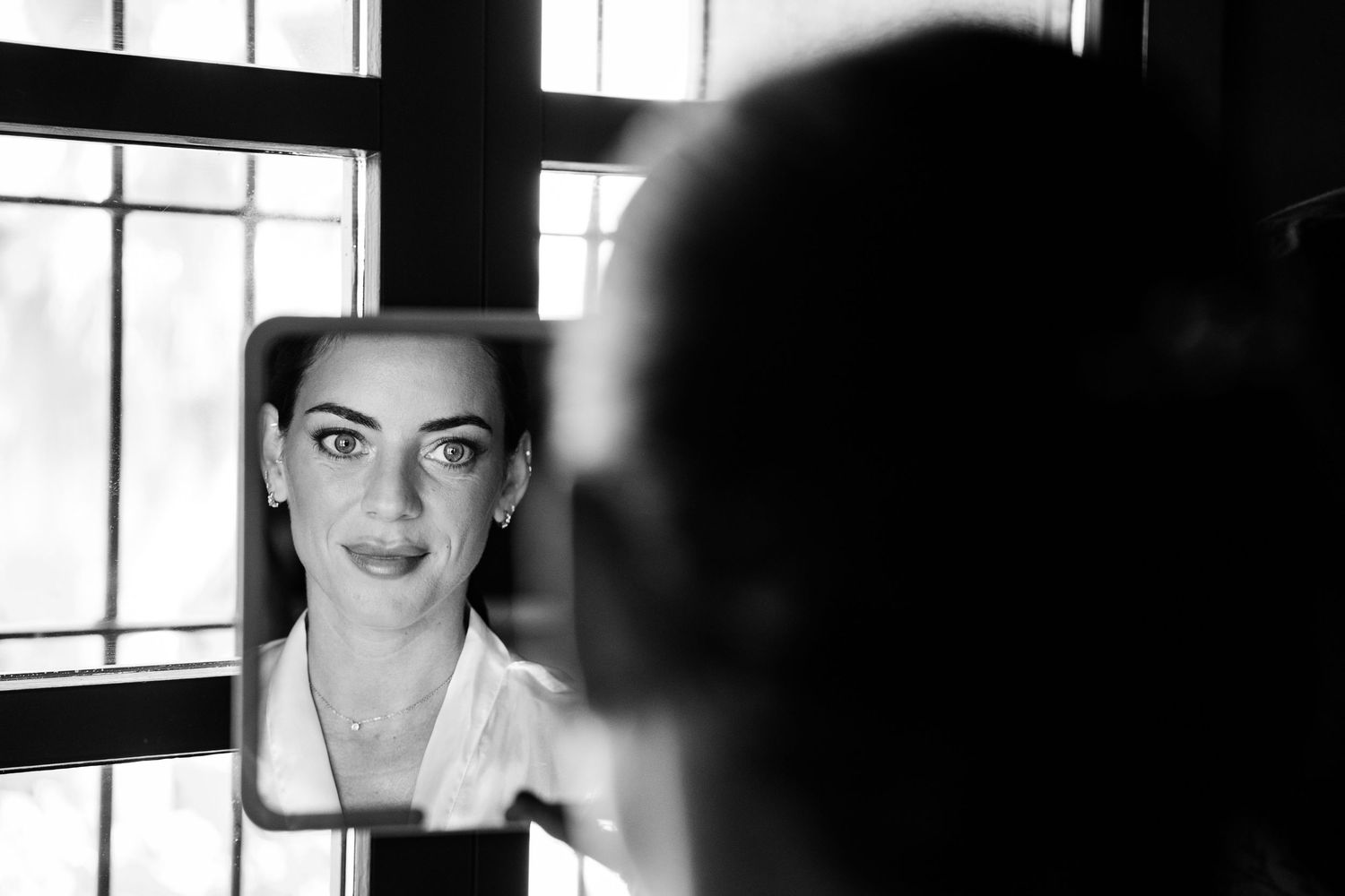 Black and white portrait of bride checking makeup in mirror before wedding ceremony.