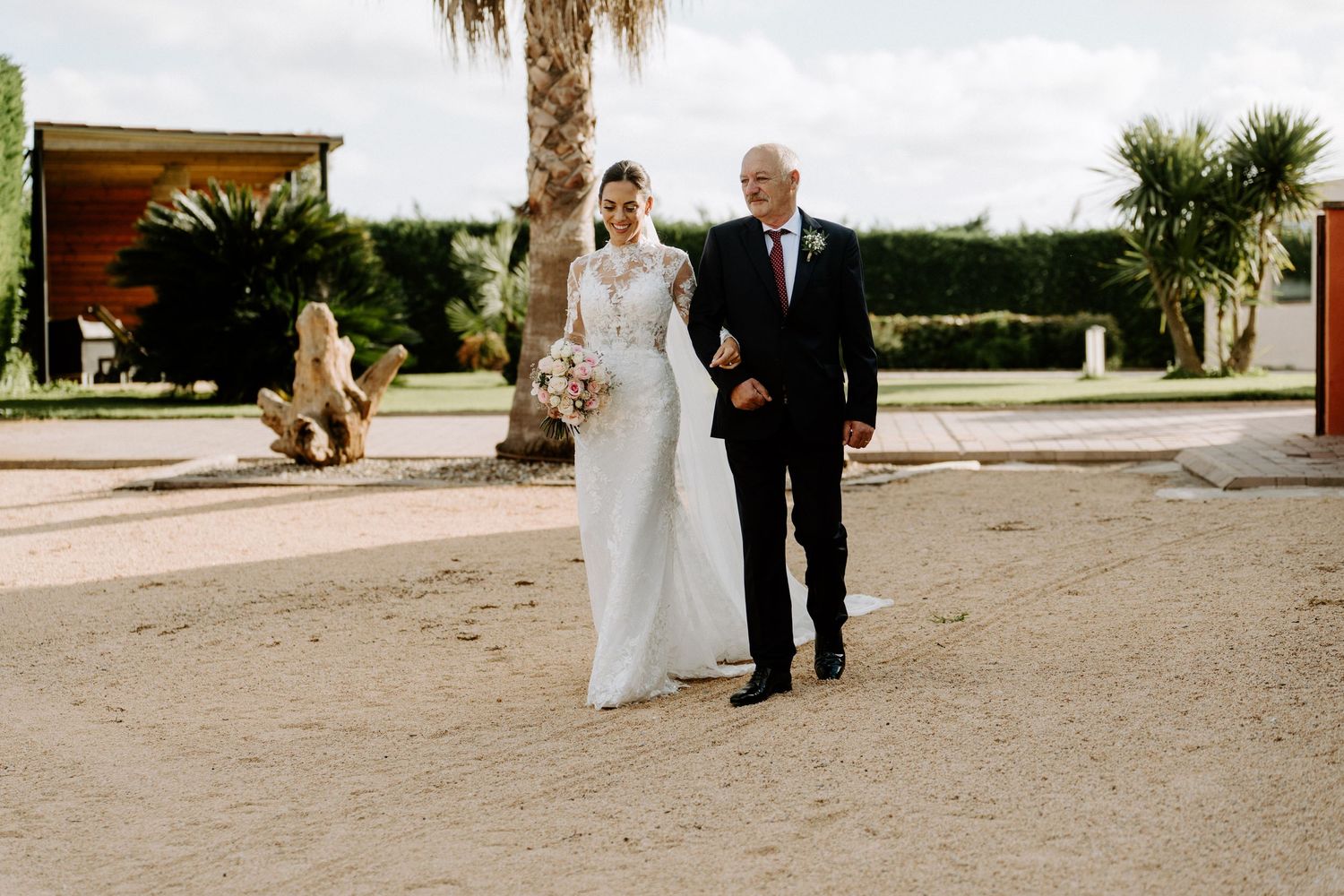 Father walks bride down sandy outdoor aisle toward wedding ceremony venue.