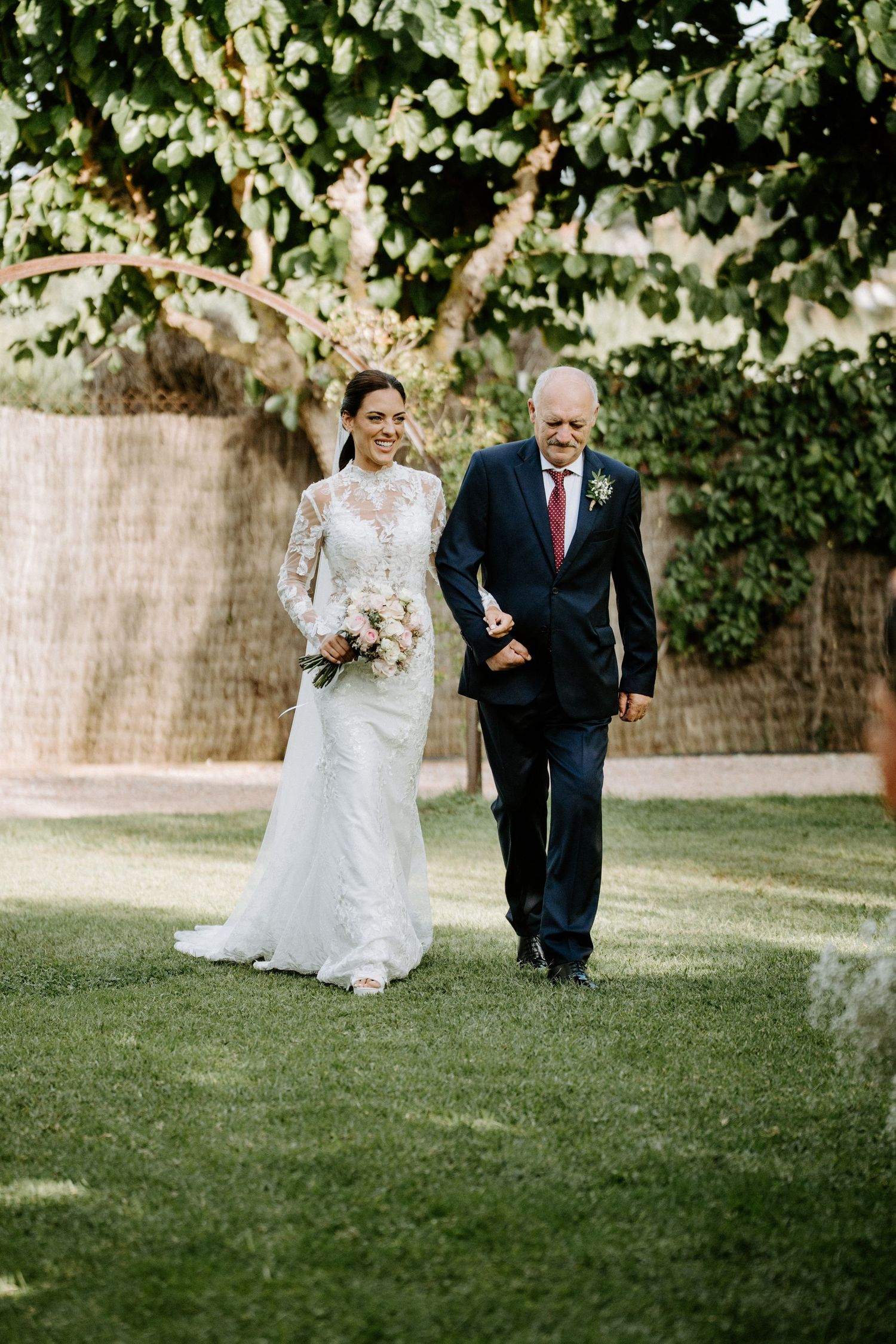 Bride in lace long-sleeve gown walks with older gentleman in dark suit through garden during outdoor wedding ceremony.