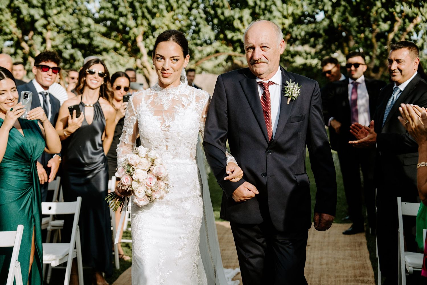 Bride in lace gown walks down aisle with father at outdoor wedding ceremony.