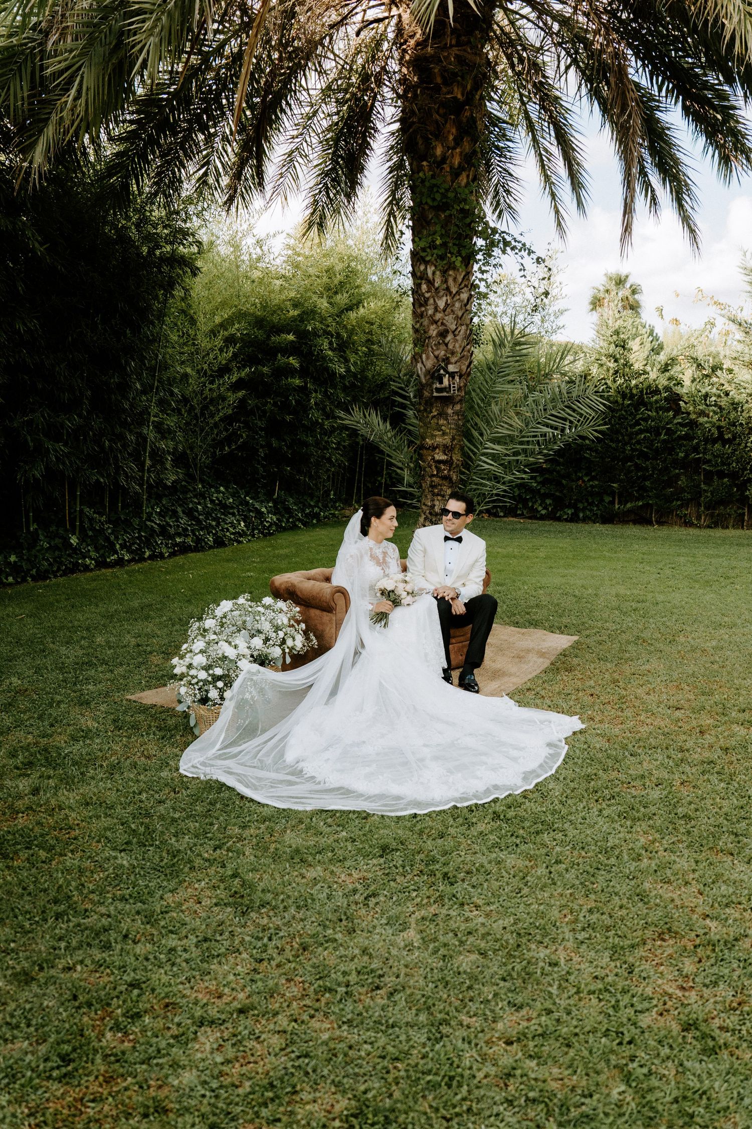 Bride and groom sitting together on a rug under a palm tree in a lush garden setting.