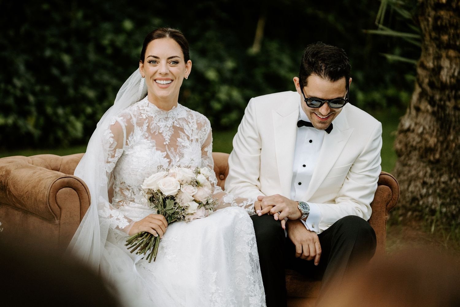 Smiling bride and groom in white tuxedo seated on vintage sofa during outdoor ceremony.