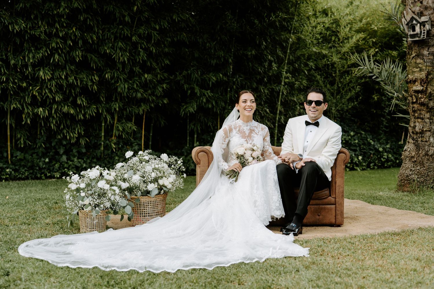 Bride and groom pose on green lawn with floral arrangements at outdoor wedding venue.