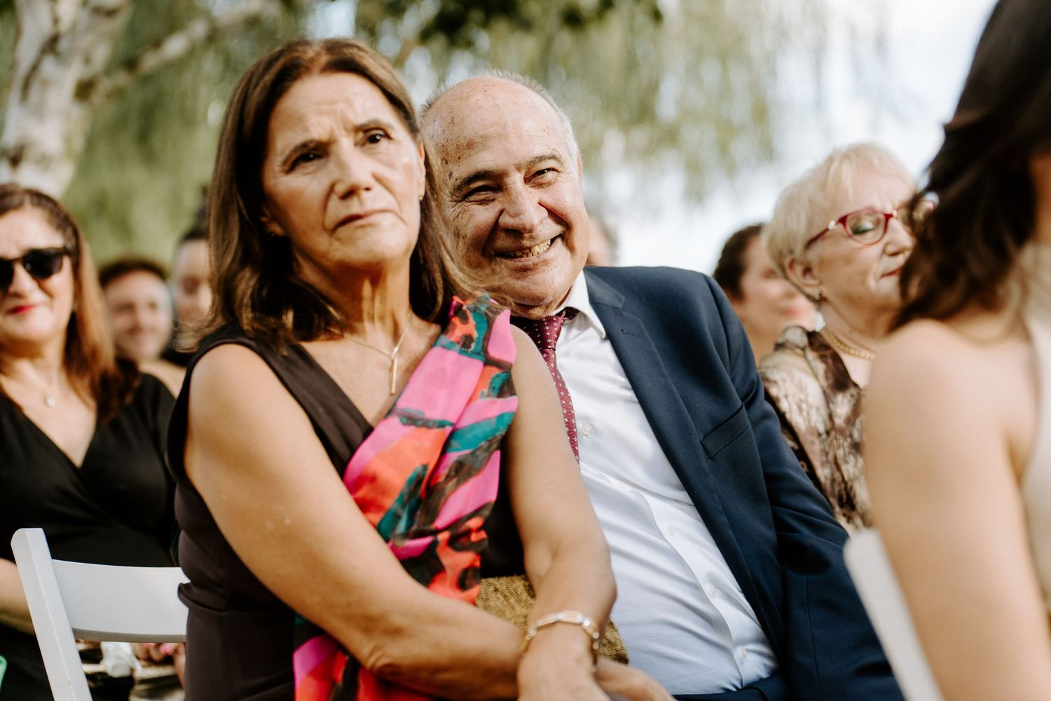Proud parents of bride smiling emotionally during outdoor wedding ceremony.