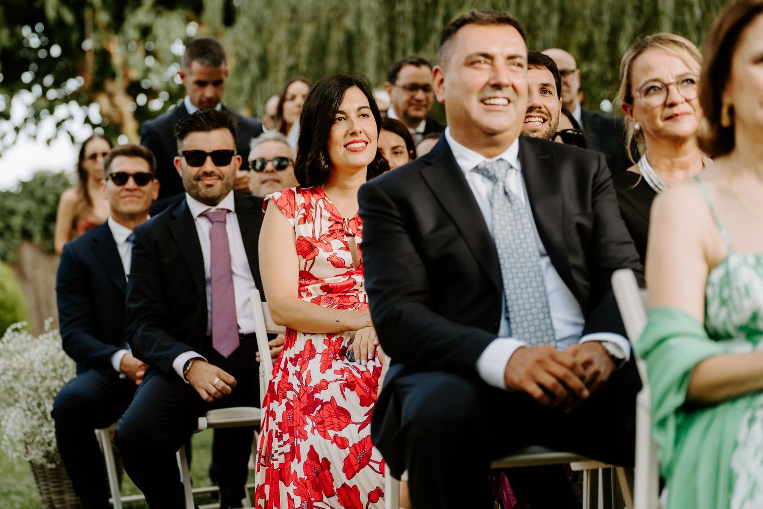 Wedding guests smiling and laughing during an outdoor ceremony seated in white chairs.