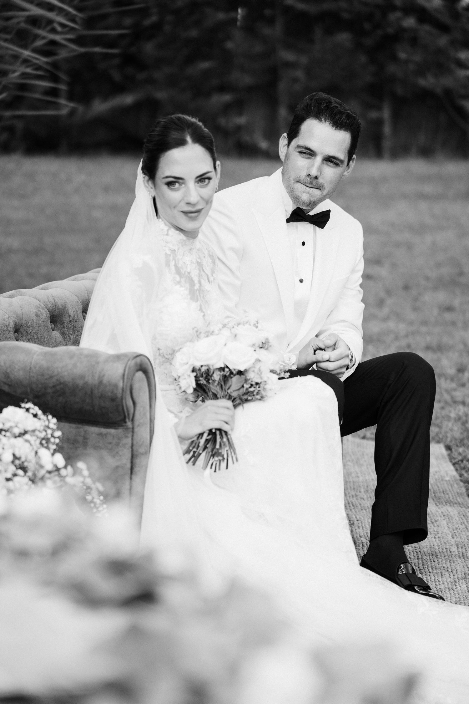 Black and white portrait of bride and groom sitting on vintage sofa holding bouquet outdoors.