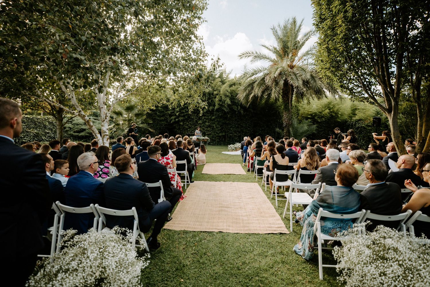 Large outdoor wedding ceremony with guests seated in rows along a floral-lined aisle.