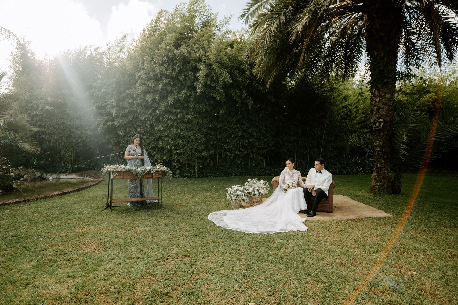 Bride and groom seated outdoors in lush garden during intimate wedding ceremony with officiant nearby.