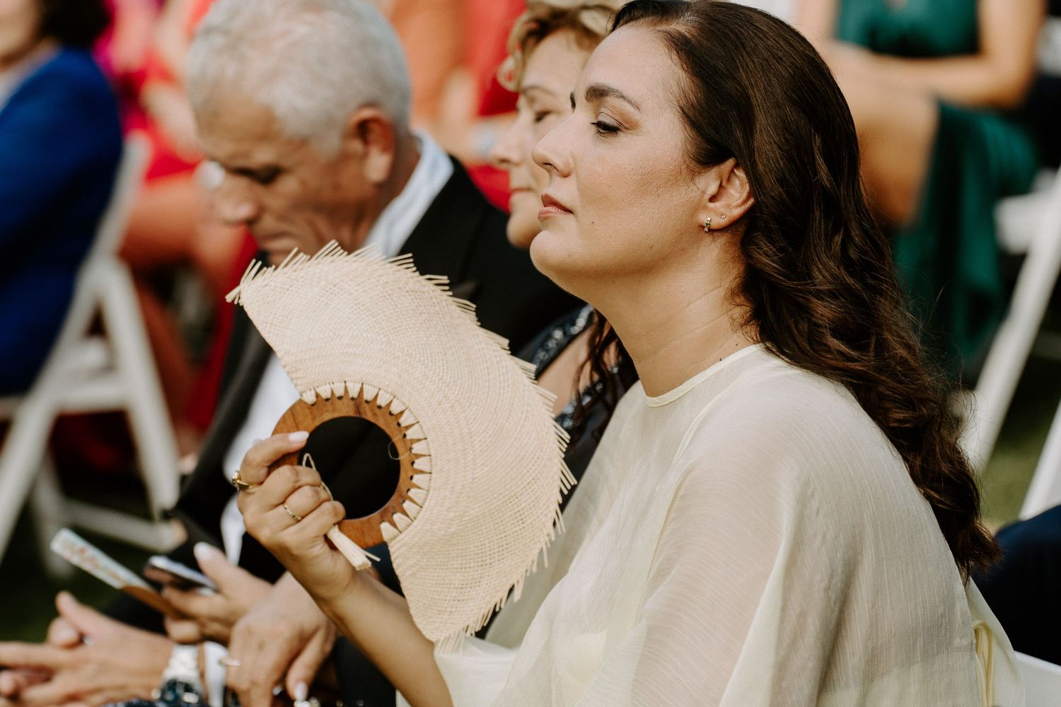 Elegant woman in white dress using a decorative fan during an outdoor wedding ceremony.