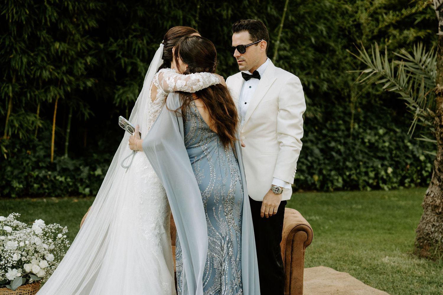 Bride in white gown and veil embraced by guests during outdoor wedding ceremony.