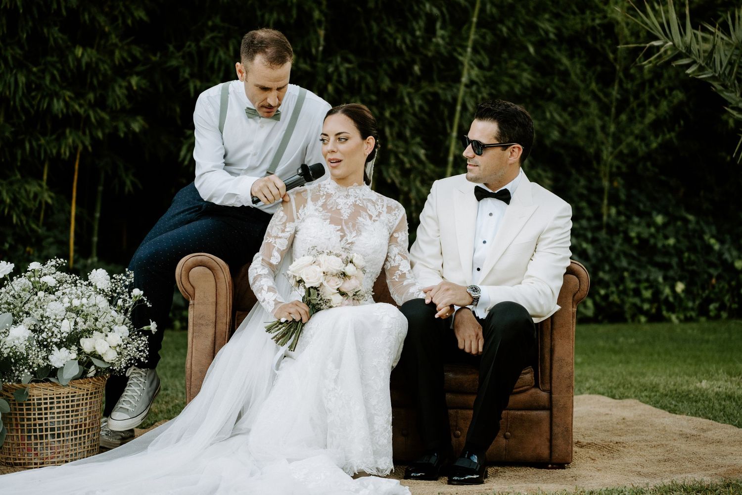 Bride in lace gown and groom in white tuxedo sitting on leather sofa with officiant outdoors.