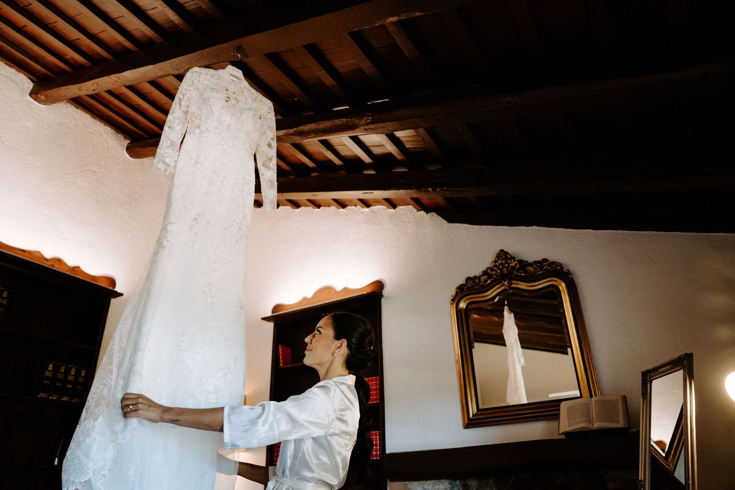 Bride admires elegant lace wedding gown hanging in rustic wooden ceiling room.