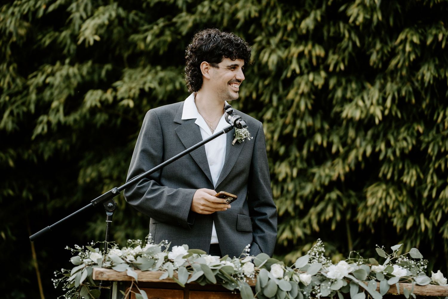 Young man in gray suit giving a speech at outdoor wedding.