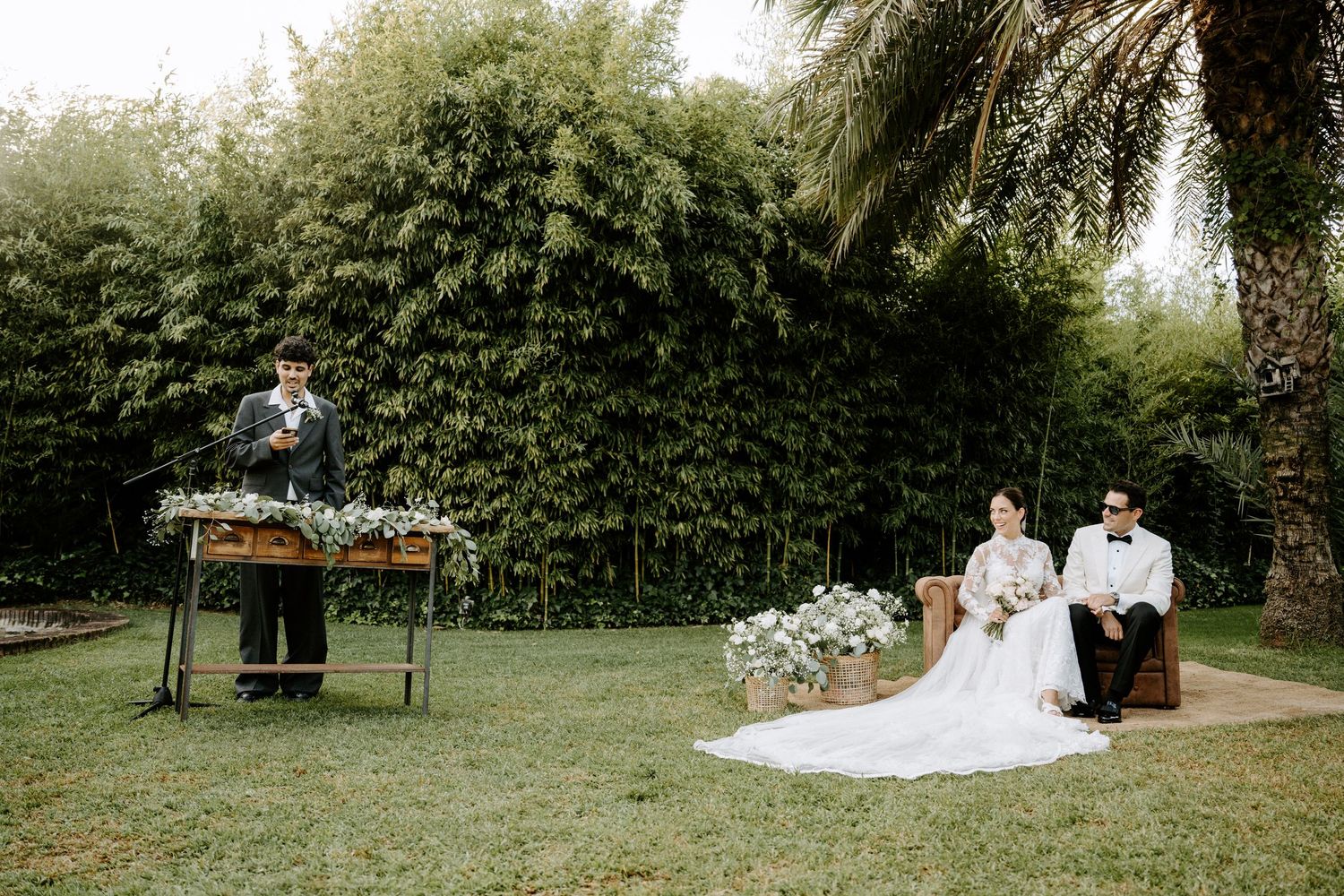 Outdoor wedding ceremony with couple seated on vintage sofa while officiant speaks at floral-decorated podium in lush garden.