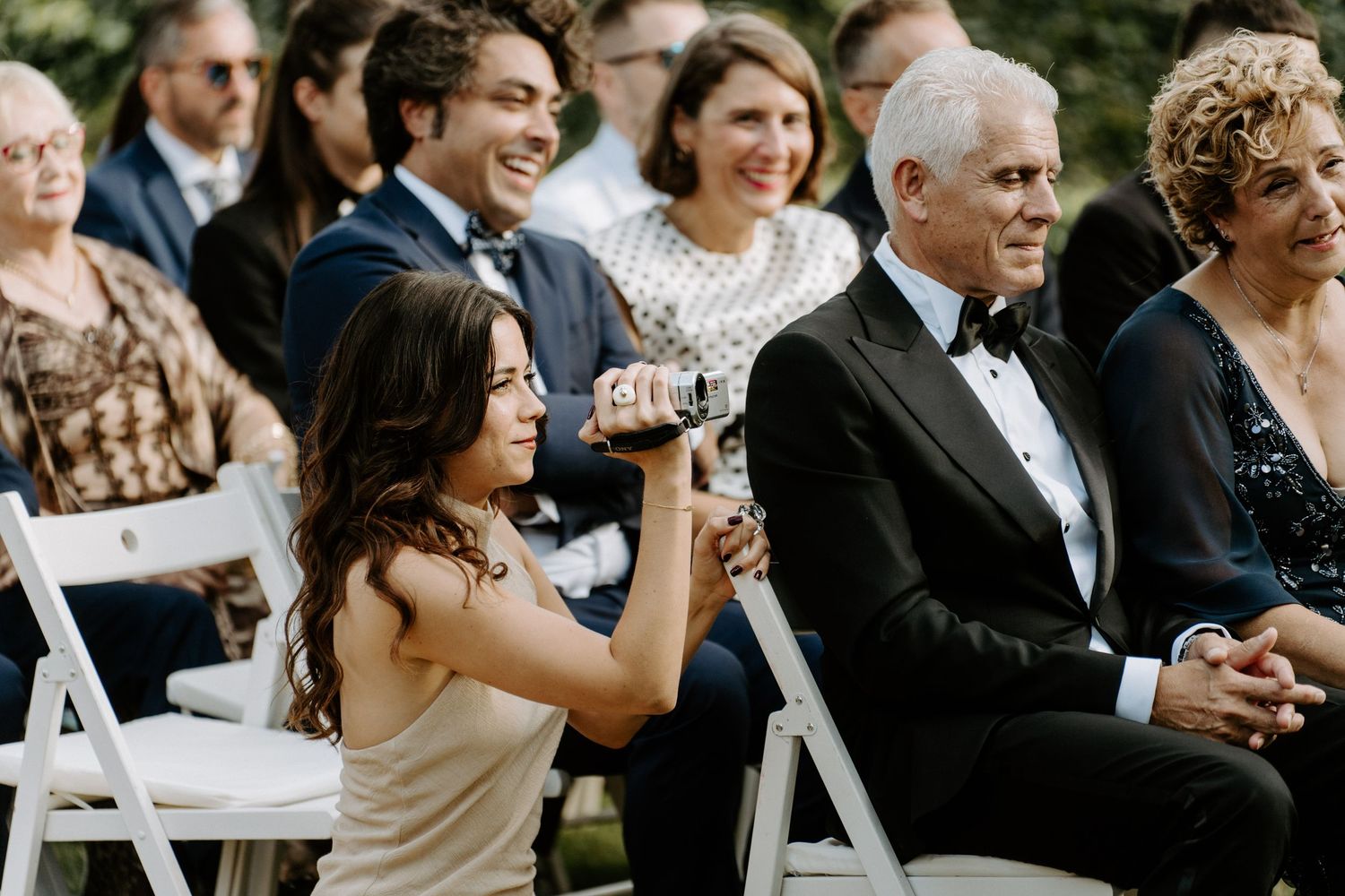 Wedding guest photographing ceremony while seated in white chairs at an outdoor wedding.