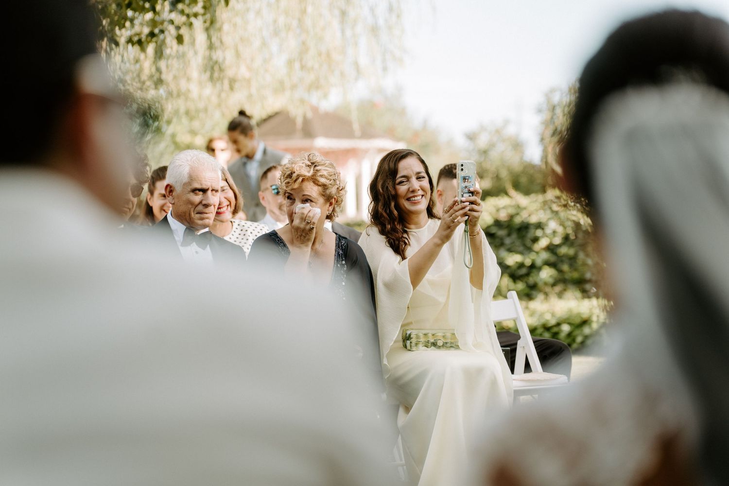 Wedding guest smiling and taking photo on smartphone during outdoor ceremony.
