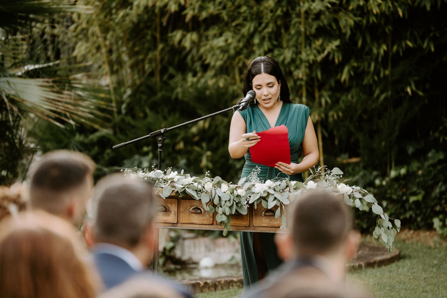 Woman in green dress reading from red book at outdoor wedding ceremony podium.