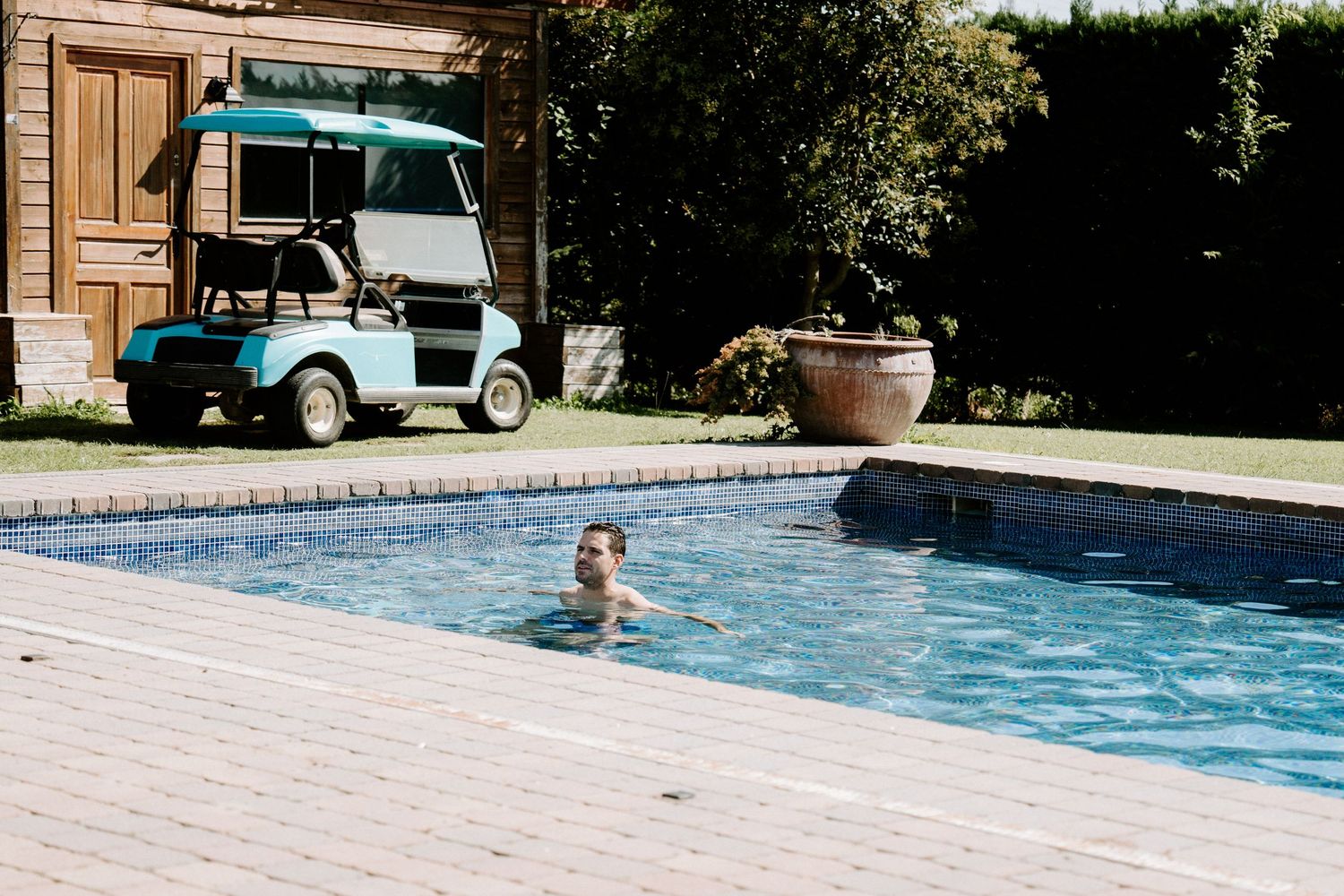 Person swimming in outdoor pool beside a golf cart and wooden building.