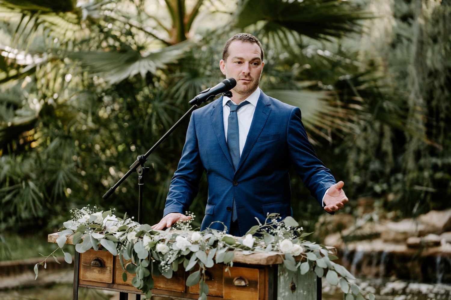 Elegant man in blue suit delivering speech at flower-adorned wooden podium during outdoor wedding ceremony.
