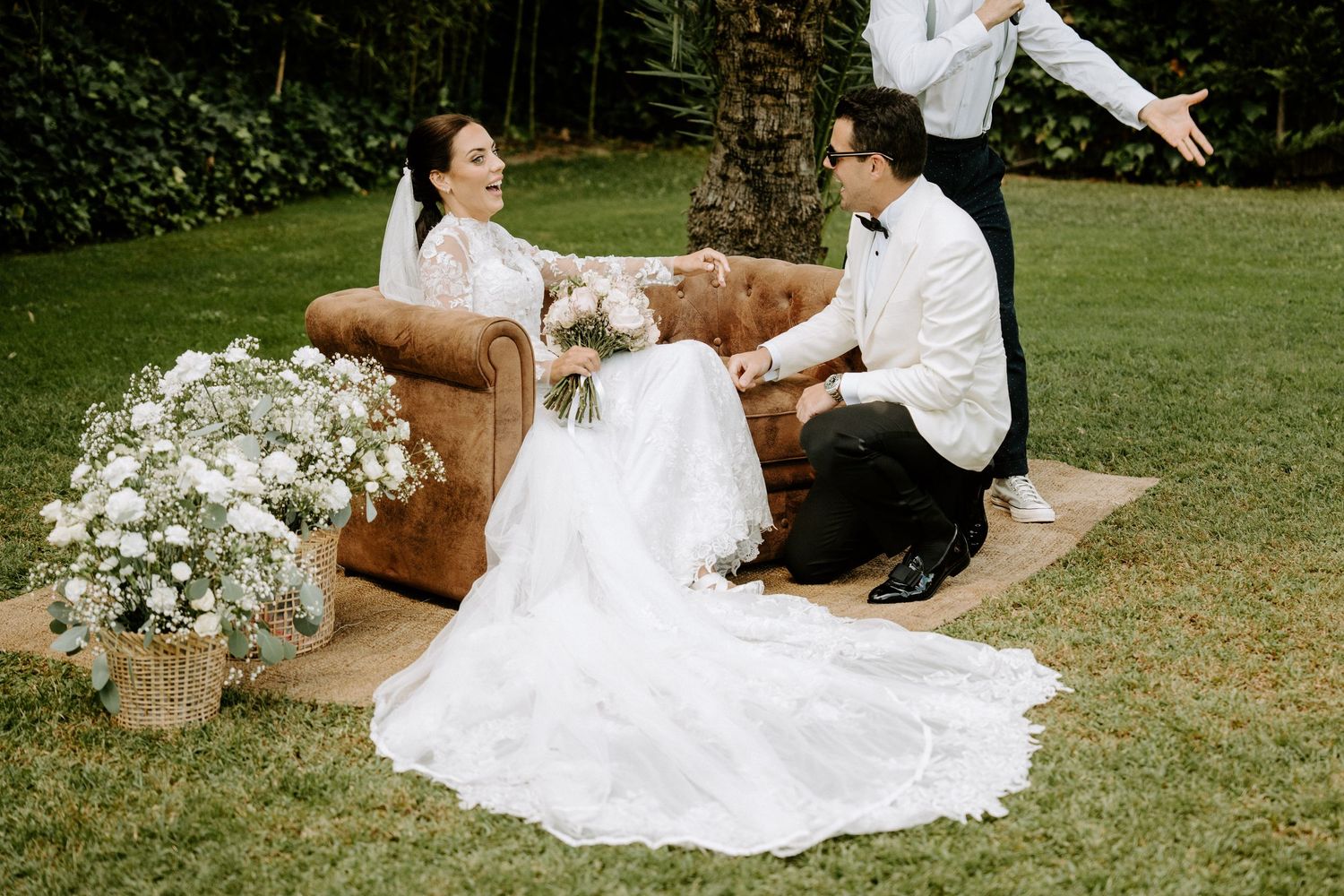 Bride in white gown and groom share joyful moment on leather sofa surrounded by white flowers at outdoor wedding.