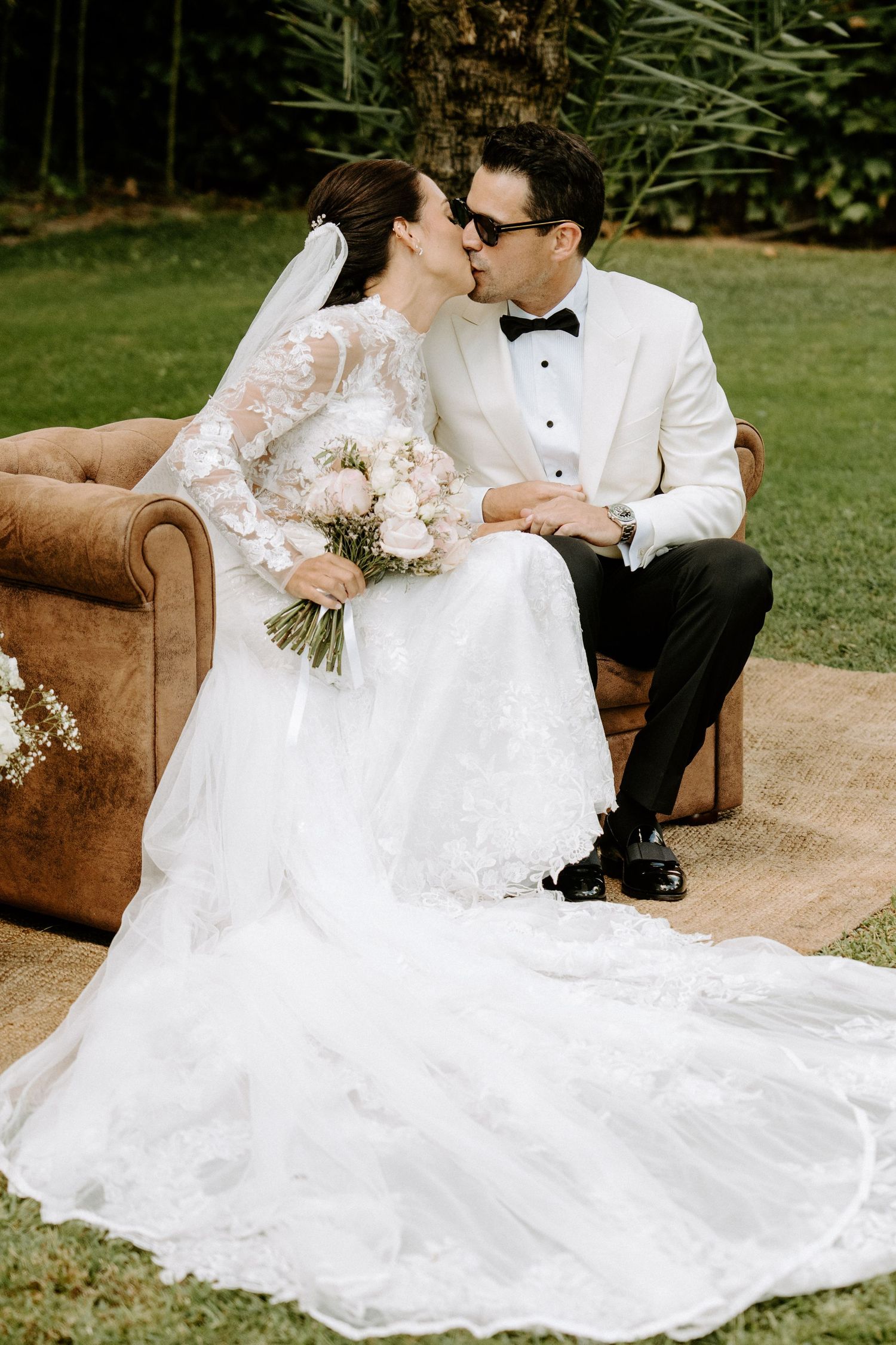 Bride and groom sharing a kiss on a leather sofa outdoors, bride holding pink bouquet.